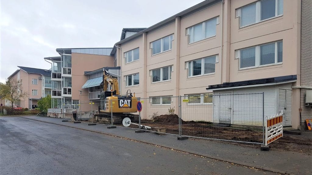 Katualue aidattuna kerrostalon edestä työmaan vuoksi. Työmaa-aitojen sisällä kaivinkone.