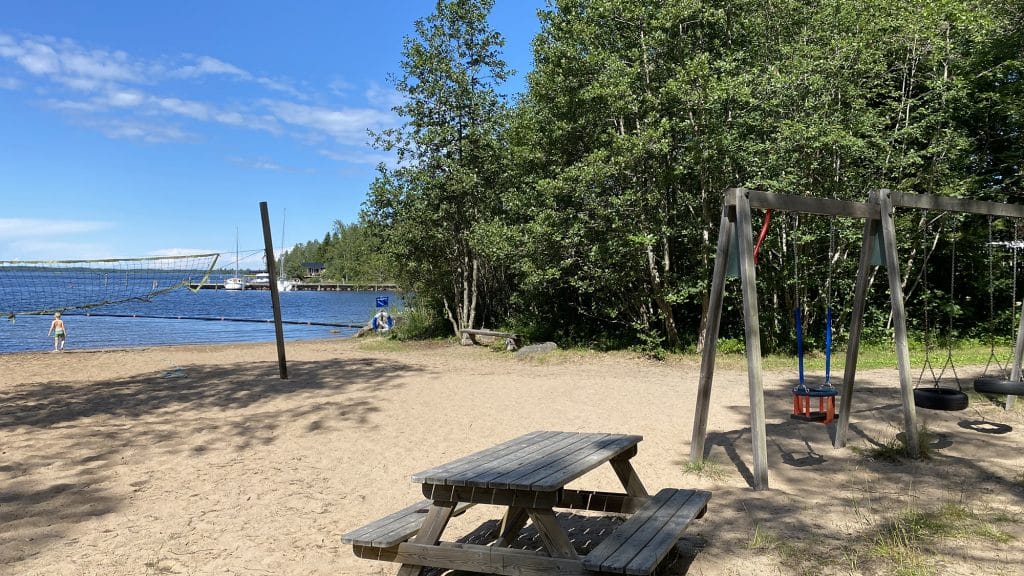Långön ranta Öjassa. Kuvan vasemmassa laidassa näkyy rantalentopallokenttä, jonka takana siintää meri ja rantavedessä kahlaava uimari. Keskellä, kuvan etualalla on puinen pöytä penkkeineen ja sen oikealla puolella puurakenteisia keinuja. Kuvan taustalla kasvaa paljon puita.