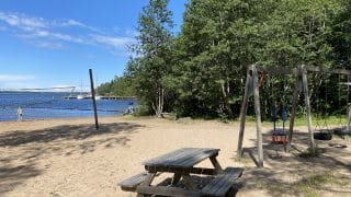 Långön ranta Öjassa. Kuvan vasemmassa laidassa näkyy rantalentopallokenttä, jonka takana siintää meri ja rantavedessä kahlaava uimari. Keskellä, kuvan etualalla on puinen pöytä penkkeineen ja sen oikealla puolella puurakenteisia keinuja. Kuvan taustalla kasvaa paljon puita.