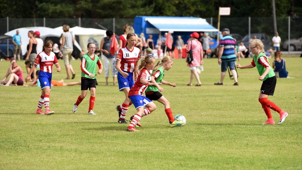 Tytöt pelaamassa jalkapalloa nurmikentällä, Kokkola Cup -turnauksessa. Kuvan keskellä vihreään liiviin ja mustiin shortseihin pukeutunut tyttö kuljettaa palloa ja hänen kannoillaan palloa tavoittelee punavalkoraidalliseen paitaan ja sinisiin shortseihin pukeutunut tyttö. Heidän ympärillään on molempien joukkueiden pelaajia ja kuvan taustalla näkyy peliä katsovia ihmisiä.