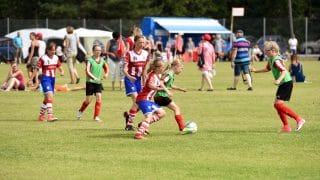 Tytöt pelaamassa jalkapalloa nurmikentällä, Kokkola Cup -turnauksessa. Kuvan keskellä vihreään liiviin ja mustiin shortseihin pukeutunut tyttö kuljettaa palloa ja hänen kannoillaan palloa tavoittelee punavalkoraidalliseen paitaan ja sinisiin shortseihin pukeutunut tyttö. Heidän ympärillään on molempien joukkueiden pelaajia ja kuvan taustalla näkyy peliä katsovia ihmisiä.