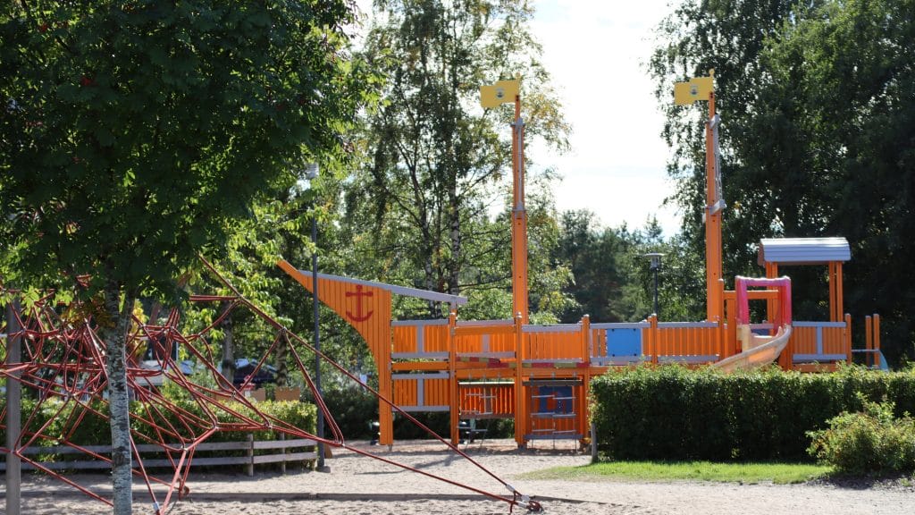 Meripuistossa oleva, puiden ympäröimä, hiekkapohjainen lasten leikkipaikka. Kuvan keskellä on laivan muotoinen kiipeilyteline liukumäkineen. Vasemmalla, kuvan etualalla näkyy köysistä koostuva kiipeilyteline.