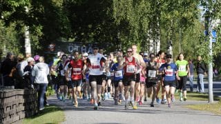 Suuri joukko juoksijoita juoksee puiden katveessa kameraa kohti. Juoksijoilla on numerolaput paidoissaan. Juoksijoiden joukossa on miehiä, naisia ja lapsia. Kuvan vasemmassa laidassa on aita, jonka takana on ihmisiä juoksua katsomassa.