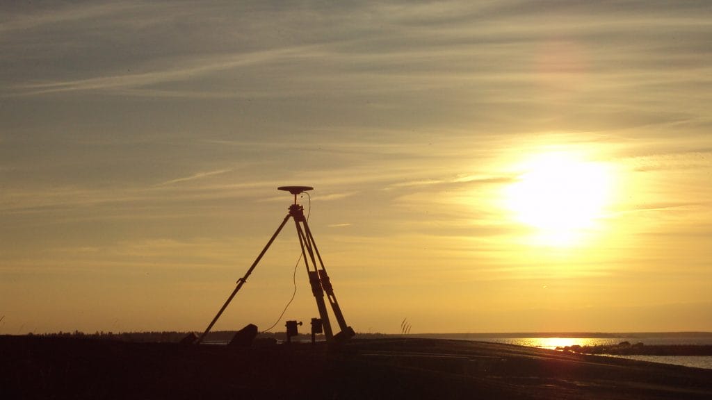 Tukipistemittausta rannalla. Hämärtyvässä illassa otettu kuva aakealle rannalle pystytetystä GPS-paikantimesta. Taustalla siintää meri ja taivaalla pilviharson lomassa laskeva aurinko.
