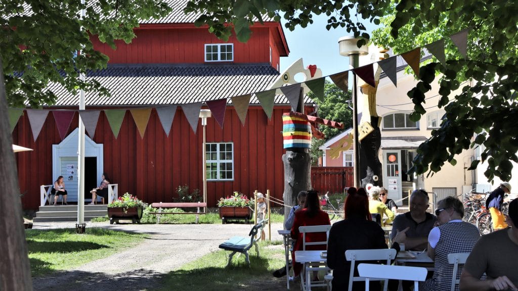 Kesäinen näkymä museopihasta. Taustalla näkyy punainen Pedagogio, jonka aukinaisten ovien molemmin puolin istuu kaksi naista tuoleillaan. Etualalla, kuvan oikeassa reunassa on joukko pöydissä istuvia ihmisiä ja heidän yläpuolellaan, puiden katveessa kulkee monivärinen viirinauha.