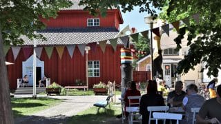 Kesäinen näkymä museopihasta. Taustalla näkyy punainen Pedagogio, jonka aukinaisten ovien molemmin puolin istuu kaksi naista tuoleillaan. Etualalla, kuvan oikeassa reunassa on joukko pöydissä istuvia ihmisiä ja heidän yläpuolellaan, puiden katveessa kulkee monivärinen viirinauha.