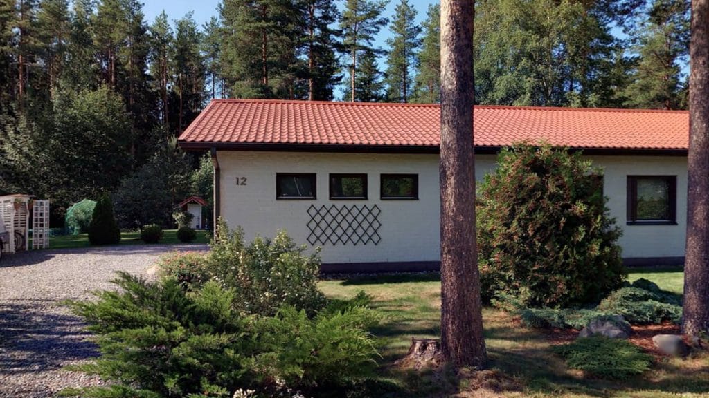 Valkoinen talo, jossa on punertava katto. Talon seinässä on osoitenumero kultaisilla numeroilla. Etualalla näkyy pihaa, jossa on pari puuta sekä erilaisia pensaita.
