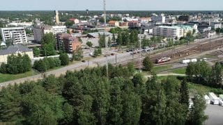 Korkealta viistosta kuvattu näkymä, jonka etualalla on puita ja niiden takaa aukeaa näkymä rautatieaseman ratapihalle sekä sen takana aukeavalle kaupungille rakennuksineen. Kaukana horisontissa näkyy myös Kokkolan vesitorni.