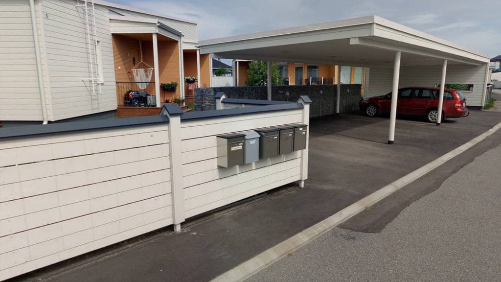 Postilaatikkorivi valko-sinisessä aidassa Asuntomessualueella. Aidan takana näkyy valkoista rakennusta, jonka kuistit on maalattu oransseiksi. Oikealla on oranssi farmariauto valkoisen autokatoksen alla.