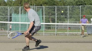 Kaksi miestä pelaamassa tennistä ulkokentällä. Lähempänä, selin kameraan olevalla miehellä on kädessään sininen maila ja yllään ruudulliset shortsit sekä harmaa t-paita. Kauempana, verkon toisella puolella olevalla miehellä on yllään lila t-paita sekä päässään lippalakki ja aurinkolasit. Verkon yläpuolella lentää keltainen tennispallo.