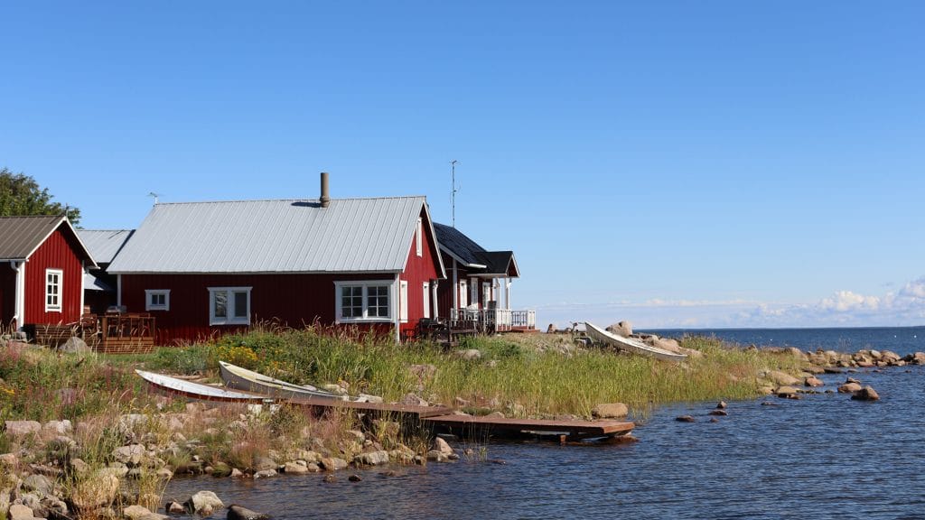 Ohtakarin kivien ja pitkän ruohon peittämä rantaviiva. Kuvan etualalla on mereen työntyvä puinen laituri sekä sen viereen maalle nostetut kaksi venettä. Taustalla näkyy punaisia, harjakattoisia rakennuksia sekä sininen, lähes pilvetön taivas. Kuvan oikeassa reunassa on tummansininen meri.