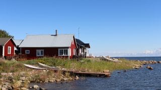 Ohtakarin kivien ja pitkän ruohon peittämä rantaviiva. Kuvan etualalla on mereen työntyvä puinen laituri sekä sen viereen maalle nostetut kaksi venettä. Taustalla näkyy punaisia, harjakattoisia rakennuksia sekä sininen, lähes pilvetön taivas. Kuvan oikeassa reunassa on tummansininen meri.