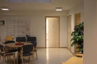 Aula, jossa takaseinällä puoliksi avonainen ovi ja sen etuvasemmalla pöytäryhmä.