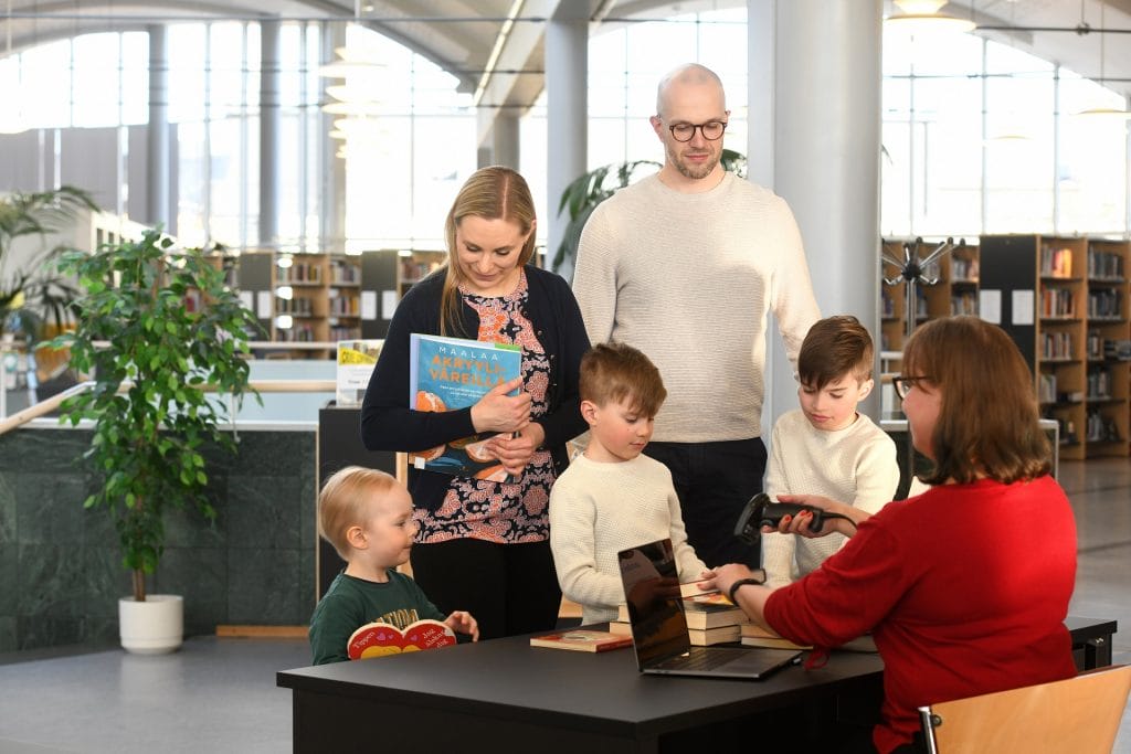 Kaksi aikuista ja kolme lasta lainaamassa kirjoja kirjastossa. Kirjastovirkailija istuu tiskin takana ja lainaa kirjat.