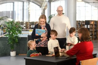 Kaksi aikuista ja kolme lasta lainaamassa kirjoja kirjastossa. Kirjastovirkailija istuu tiskin takana ja lainaa kirjat.
