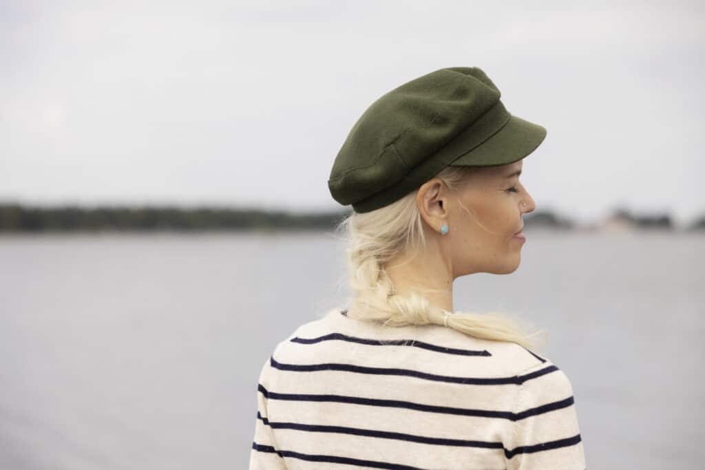Vihreää lakkiin ja mustavalkoraidalliseen paitaan pukeutunut nainen seisoo meren äärellä. selin kuvaajaan. Naisella on pitkä vaalea letti, hän hymyilee silmät suljettuina.