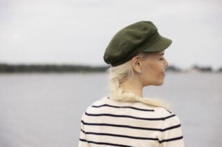 Vihreää lakkiin ja mustavalkoraidalliseen paitaan pukeutunut nainen seisoo meren äärellä. selin kuvaajaan. Naisella on pitkä vaalea letti, hän hymyilee silmät suljettuina.