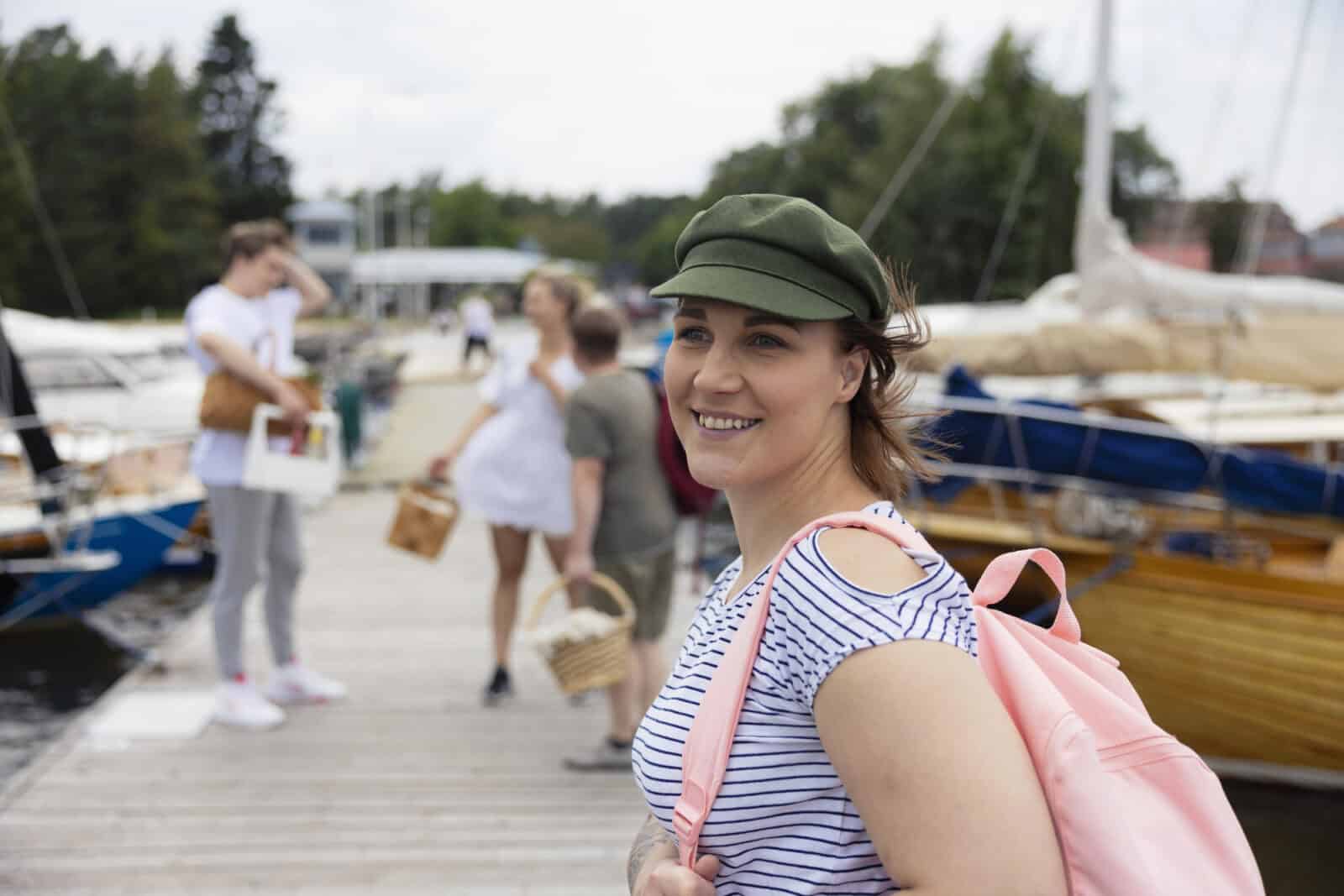 En ung kvinna som bär en grön keps står på plattformen med en rosa ryggsäck på axeln. I bakgrunden kan man se båtar och människor med lunchkorgar i händerna.