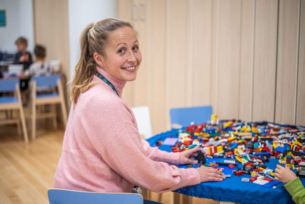 Varhaiskasvatuksen opettaja istuu askartelupöydän edessä, ja katsoo hymyillen kameraan olkansa yli.