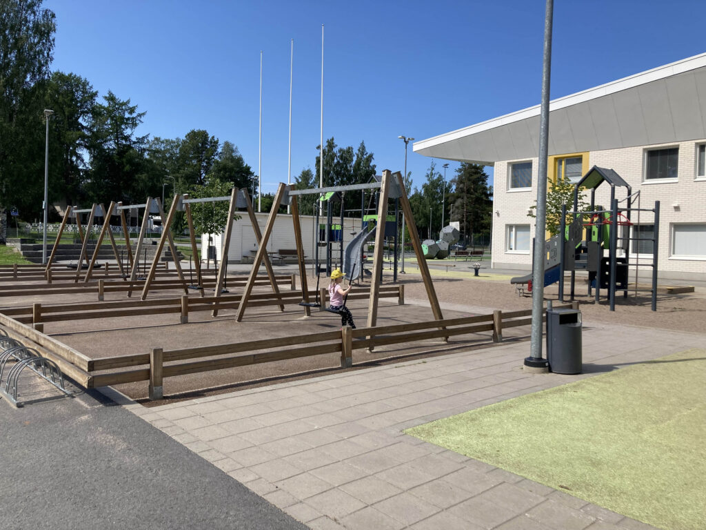 Aurinkoinen koulun piha, jonka leikkipihalla on lapsi keinumassa.